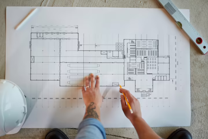 Manos marcando con lápiz un plano arquitectónico impreso junto a un casco y un nivel